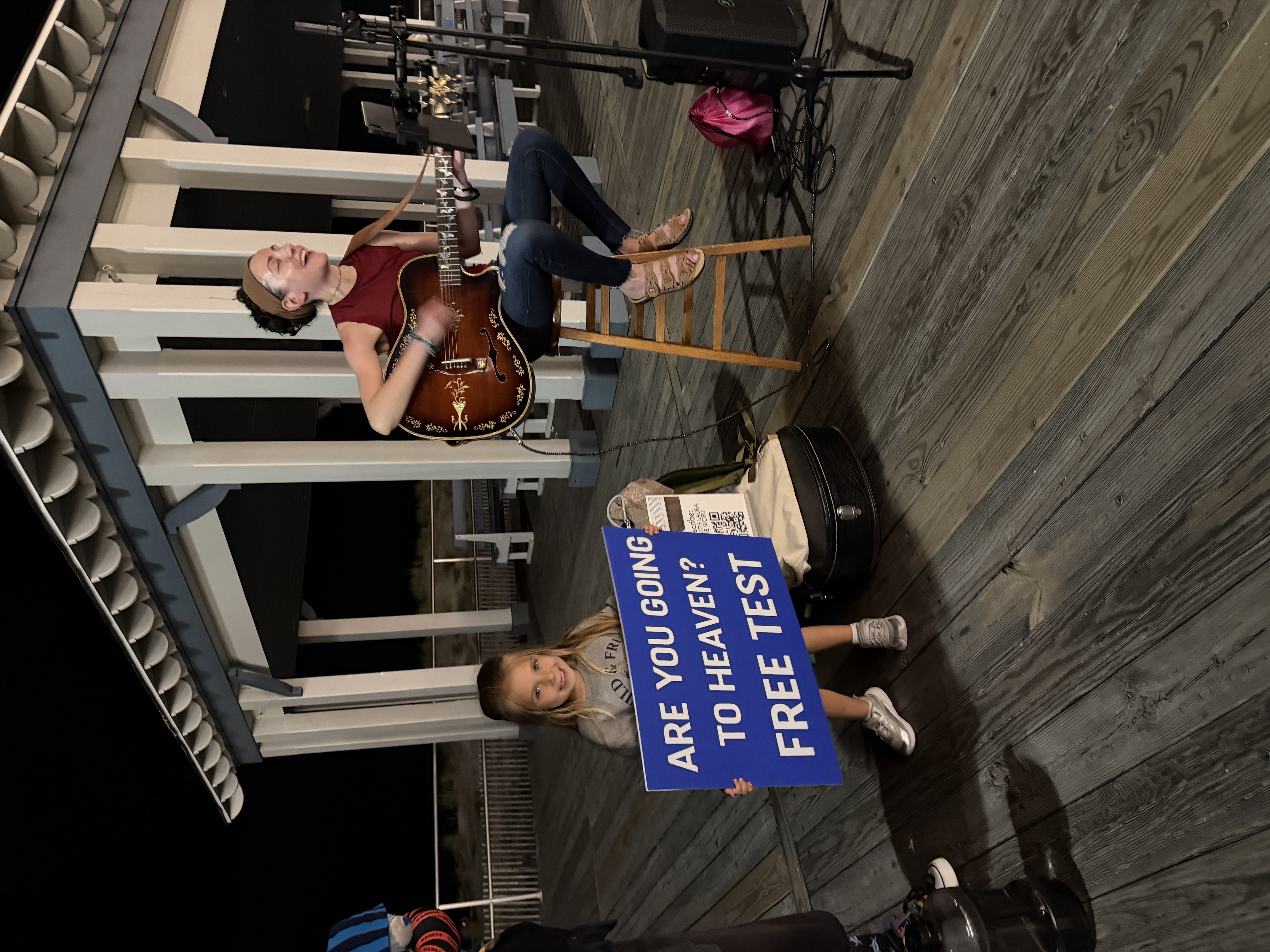 On the boardwalk next to a pavilion at nighttime, Laura singing and playing the car, standing beside her is her 3 year old granddaughter holding up a sign that says,