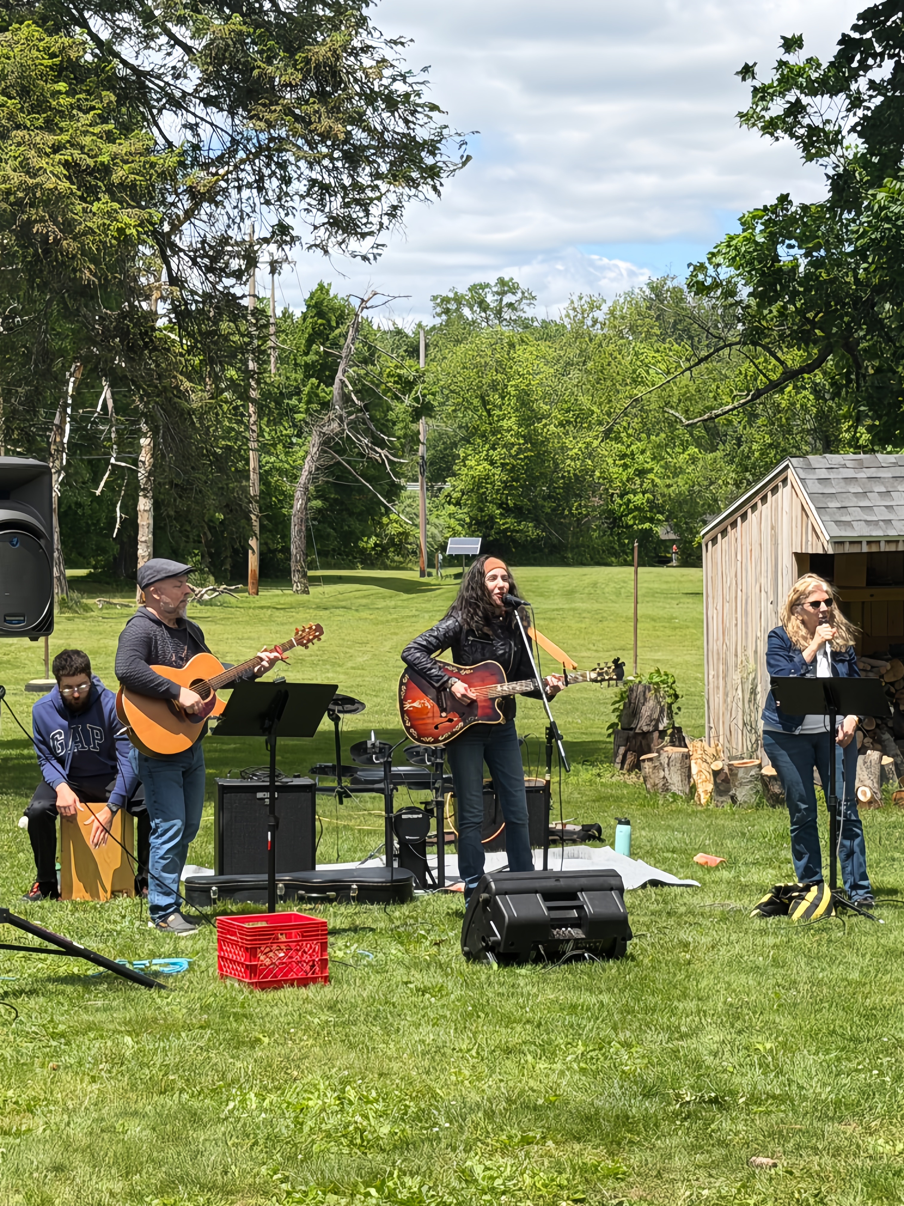 Laura and her band singing and playing guitars and a cajon box drum on grass with trees behind them at Crossing Community Church, Newtown, PA