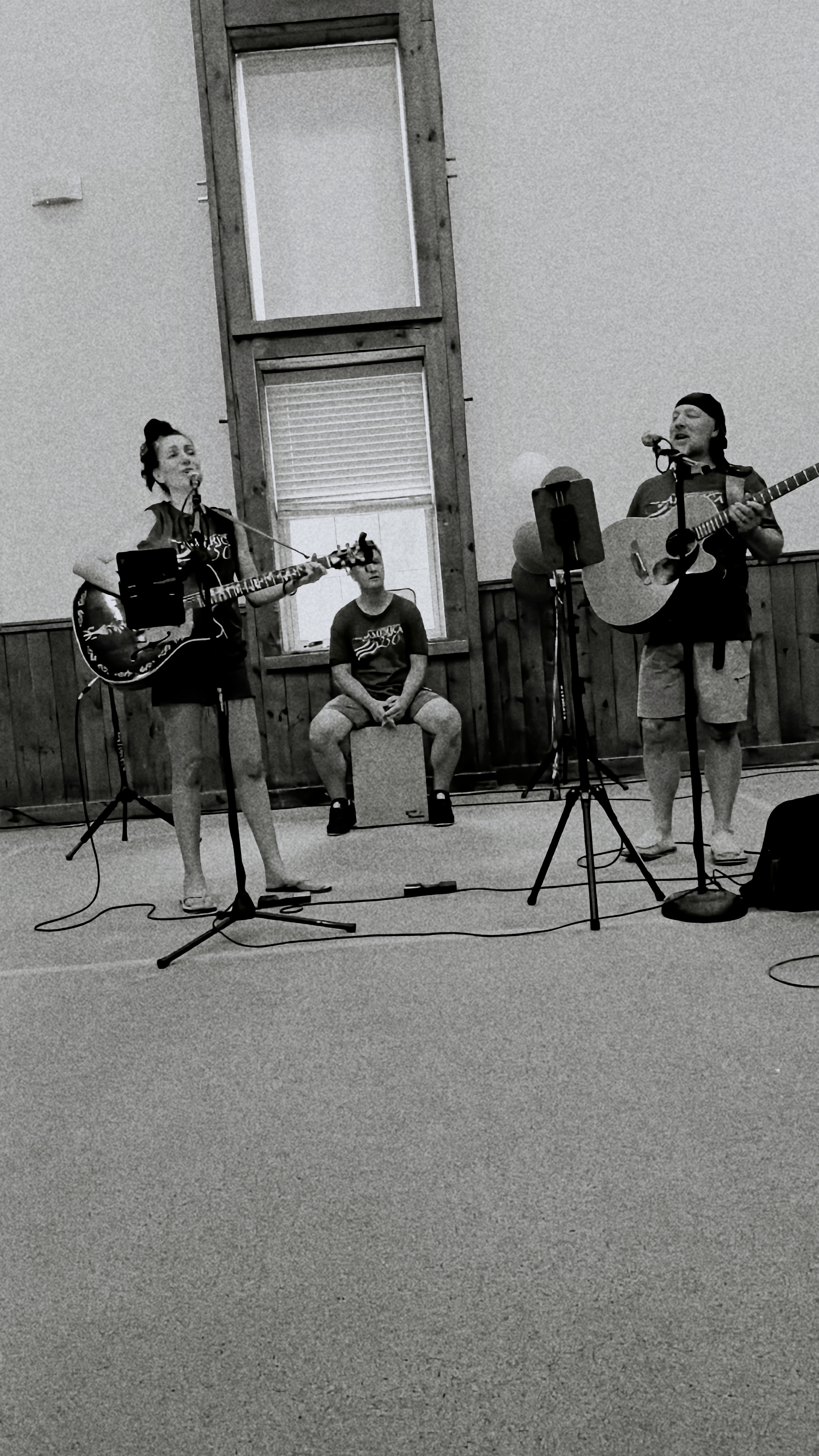 a grainy black and white photo of Laura and Michael Breece each playing guitars and singing worshipfully to God with Matt Standeven sitting on a box drum called a cajon singing also