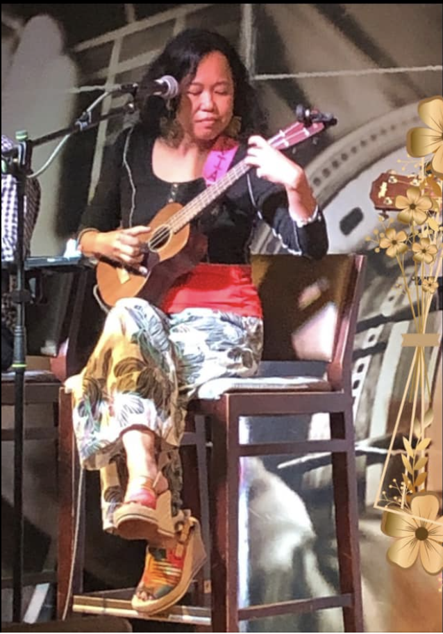 Silvia Passiflora, Southern Gothic Folk poet-songwriter seated onstage with ukulele, wearing black sweater and black and white palazzo pants