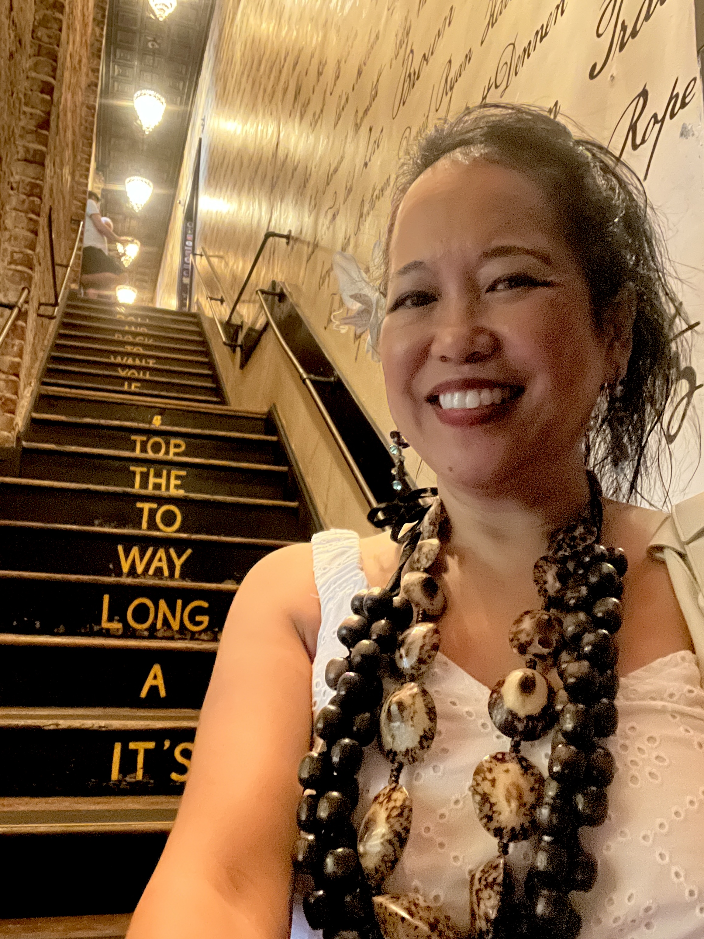 Silvia Passiflora, Southern Gothic Folk poet-songwriter. Silvia Passiflora smiling at the camera while standing in front of a staircase with song lyrics painted on the steps, wearing a bold layered statement necklace of dark beads and shell medallions and a white eyelet top.