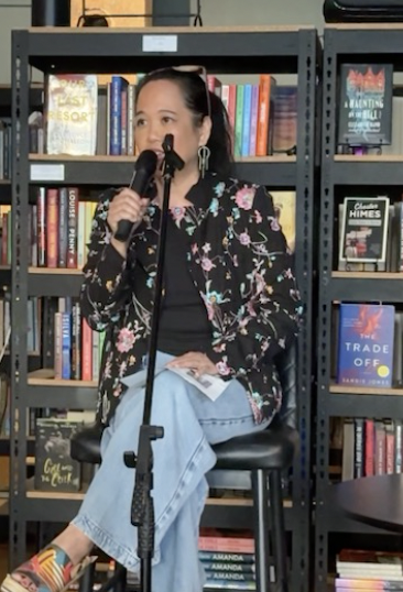 Silvia Passiflora, Southern Gothic Folk poet-songwriter, seated with a microphone in hand in front of bookshelves, wearing a floral black jacket and jeans.