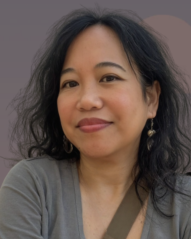 Silvia Passiflora, a Southern Gothic Folk poet-songwriter. Silvia Passiflora in a close portrait, wearing gold hoop earrings and small drop earrings, with dark wavy hair, a warm half-smile, and a gray v-neck top, against a soft gray background with a subtle circular shape behind her. 🌸