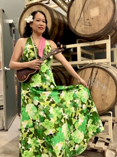 Silvia Passiflora holding an ukulele in front of wine barrels, wearing a botanical gown — Southern Gothic Folk poet-songwriter and certified sommelier.