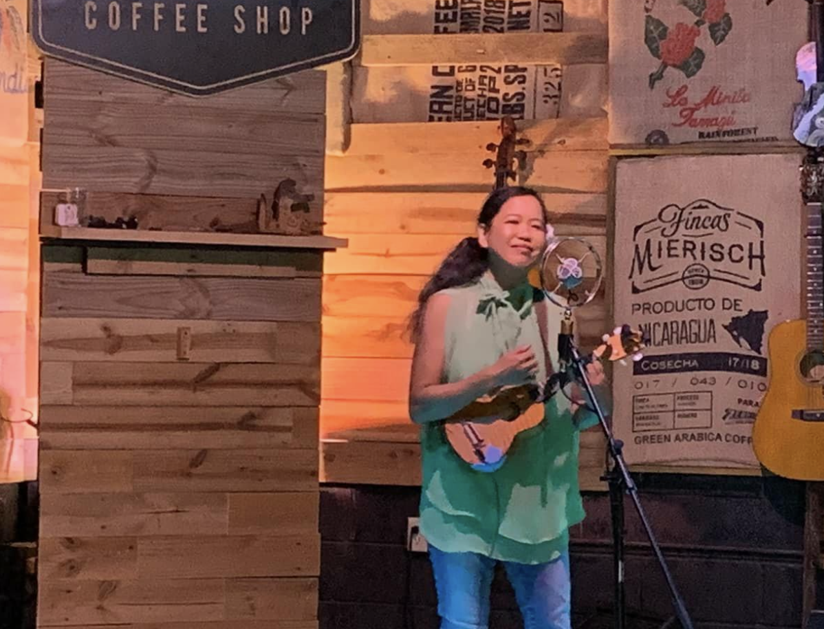Silvia Passiflora, Southern Gothic Folk poet-songwriter playing at a coffeeshop Open Mic, wearing a silk green top playing ukulele, behind a classic condenser mic.