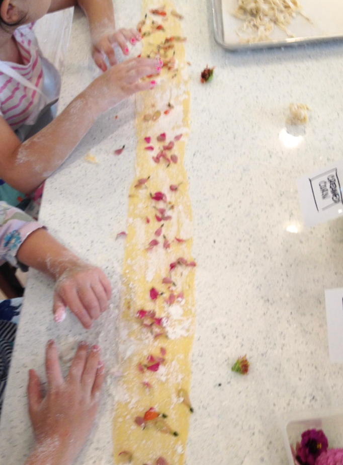 Scriptaluna — Word of Mouth Cooking Club preschool cooking class, a child's hands pressing organic flower petals into fresh pasta dough.