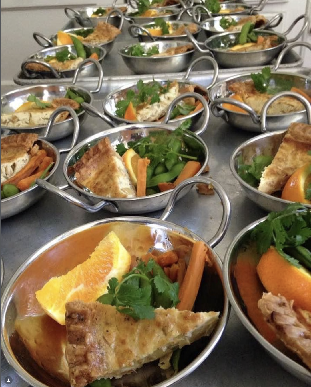 Scriptaluna — Word of Mouth Cooking Club catering, rows of stainless steel balti bowls plated with herb-crusted fish, roasted carrots, snap peas, orange slices, and fresh cilantro.