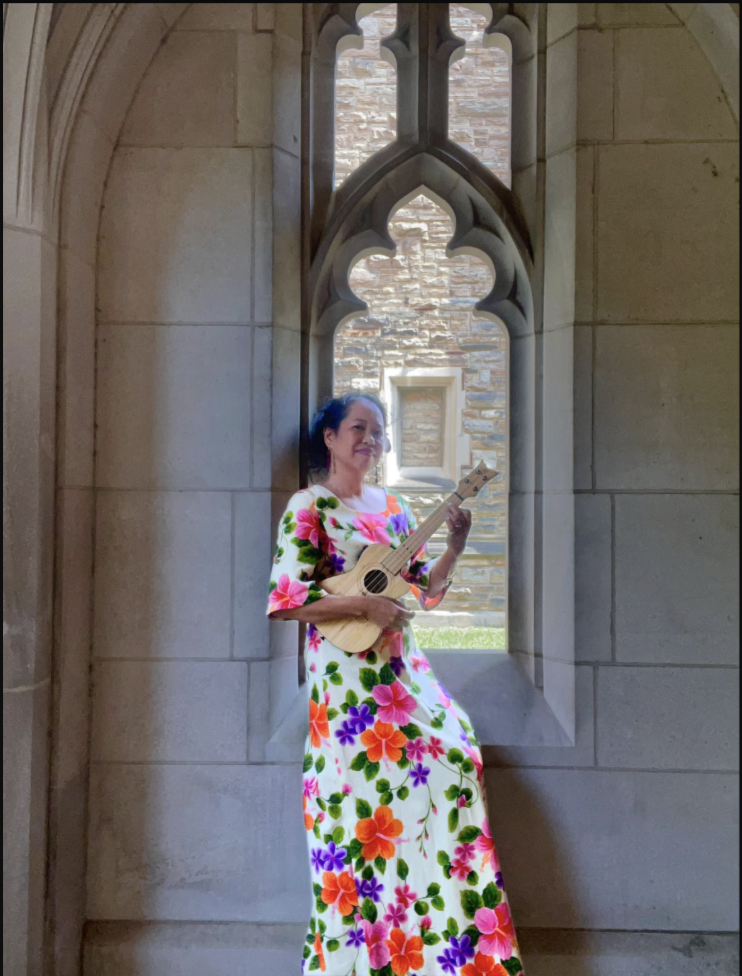 Silvia Passiflora, Southern Gothic Folk poet-songwriter wearing Hawaiiana gown holding ukulele in front of a medieval cut out window at masonry wall.