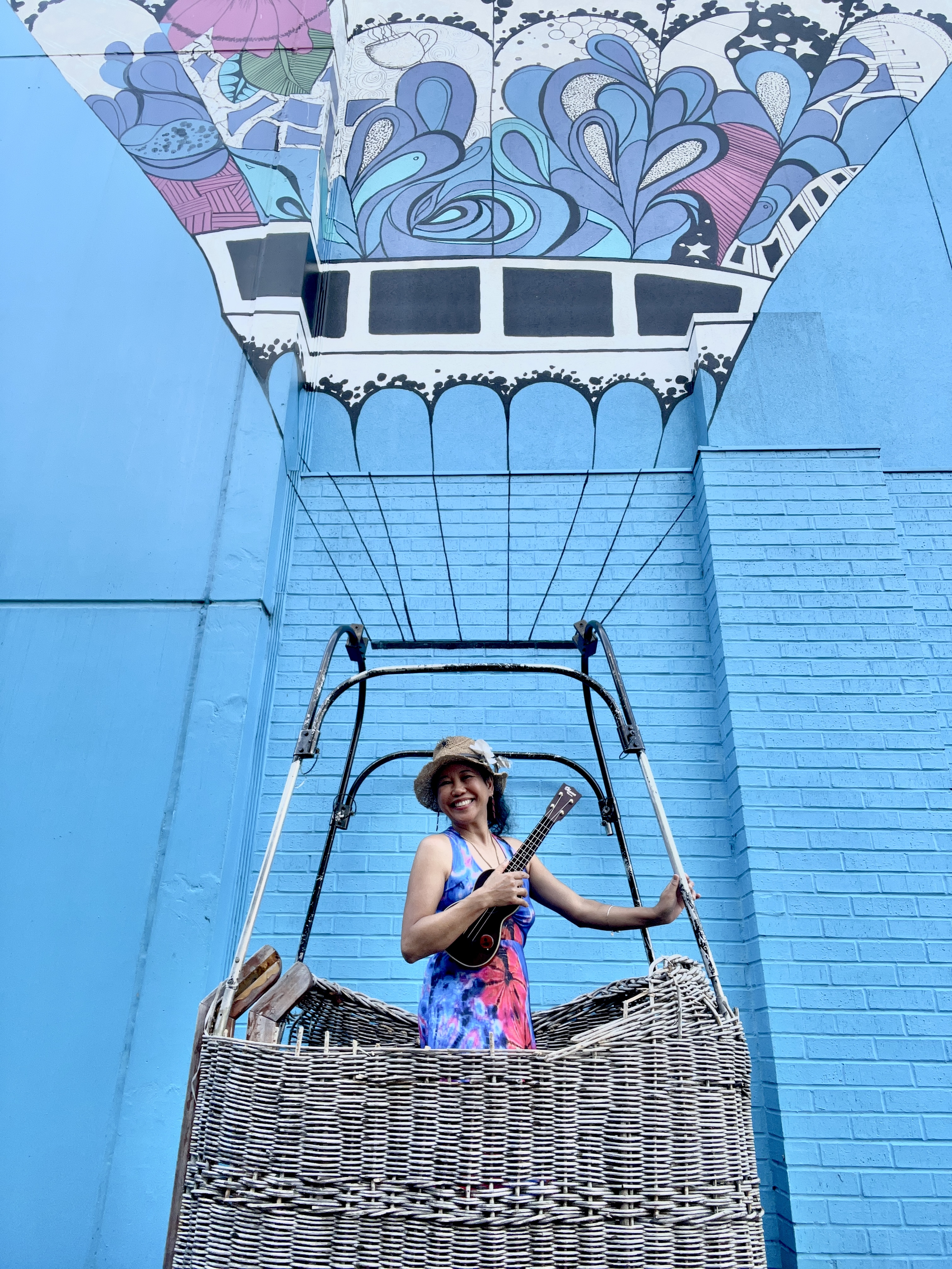 Silvia Passiflora, Southern Gothic Folk poet-songwriter. Silvia Passiflora standing in a wicker hot air balloon basket mounted against a vivid blue brick wall mural, holding an ukulele, wearing a tie-dye dress, a straw hat with a white flower, and smiling broadly upward.