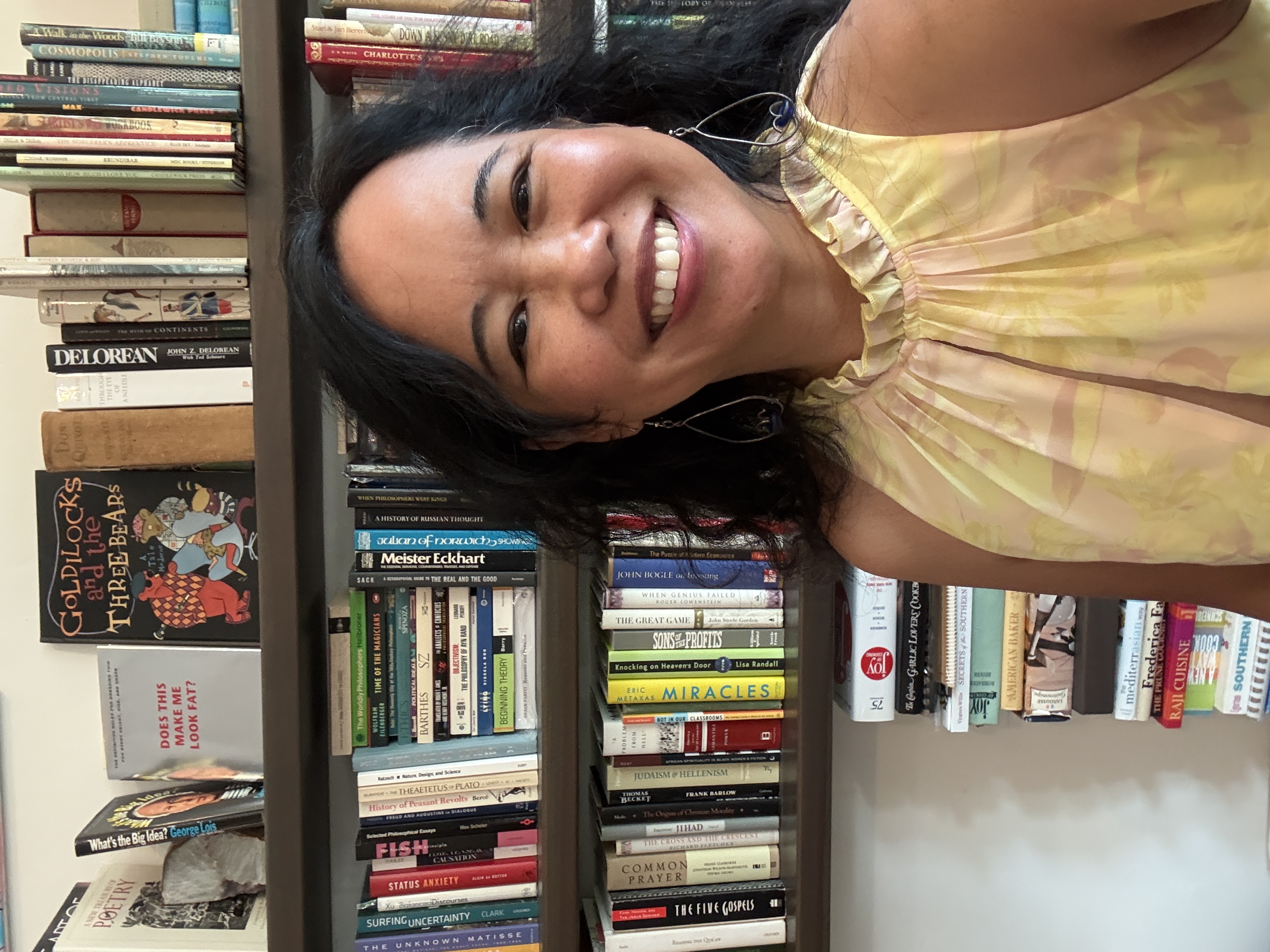 Silvia Passiflora, Southern Gothic Folk poet-songwriter and publisher of Scriptaluna, in front of books in a bookstore, wearing a buttercup yellow dress, smiling to the camera.