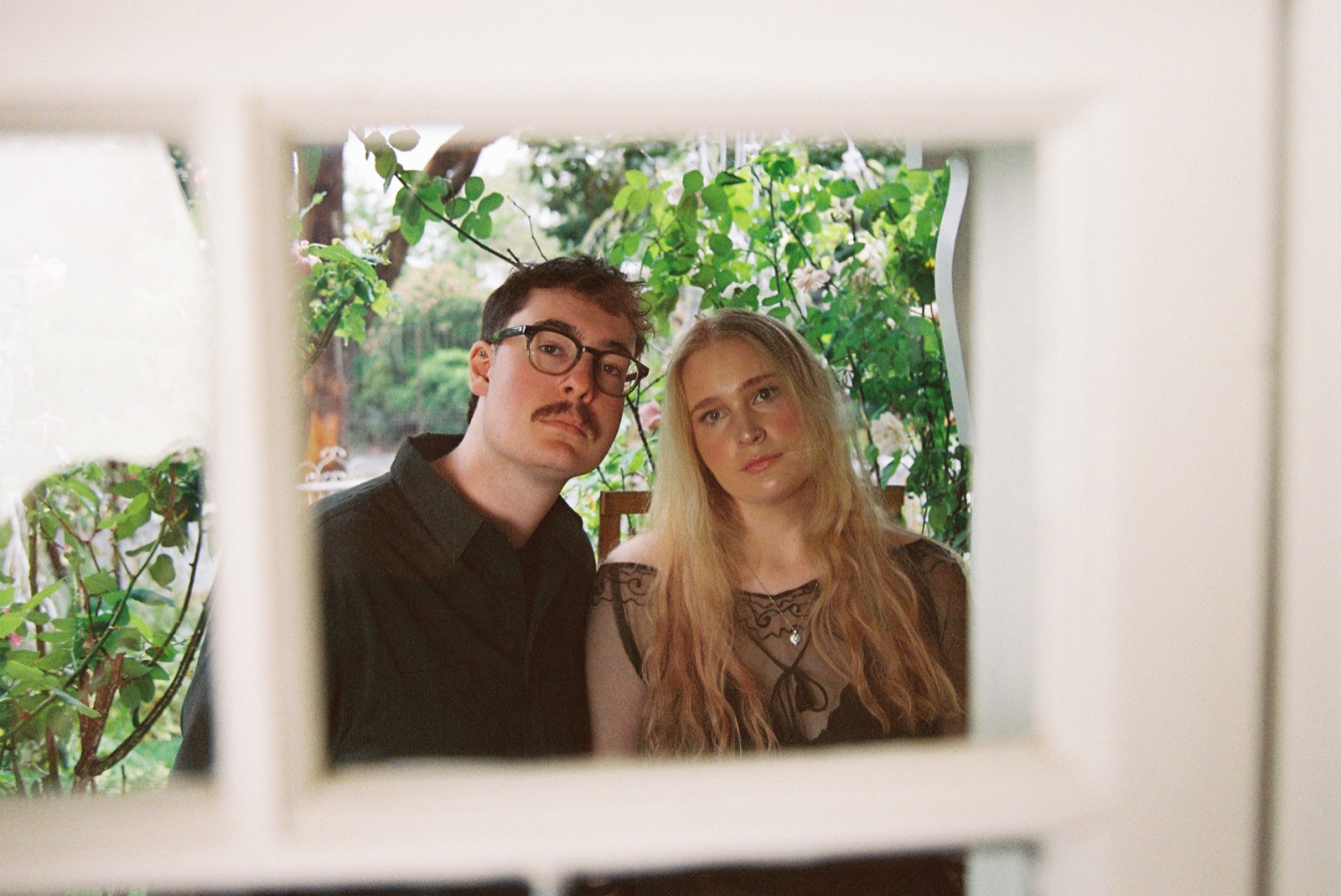 Clancy and Gabrielle McCue looking in through a white window frame, garden behind them.