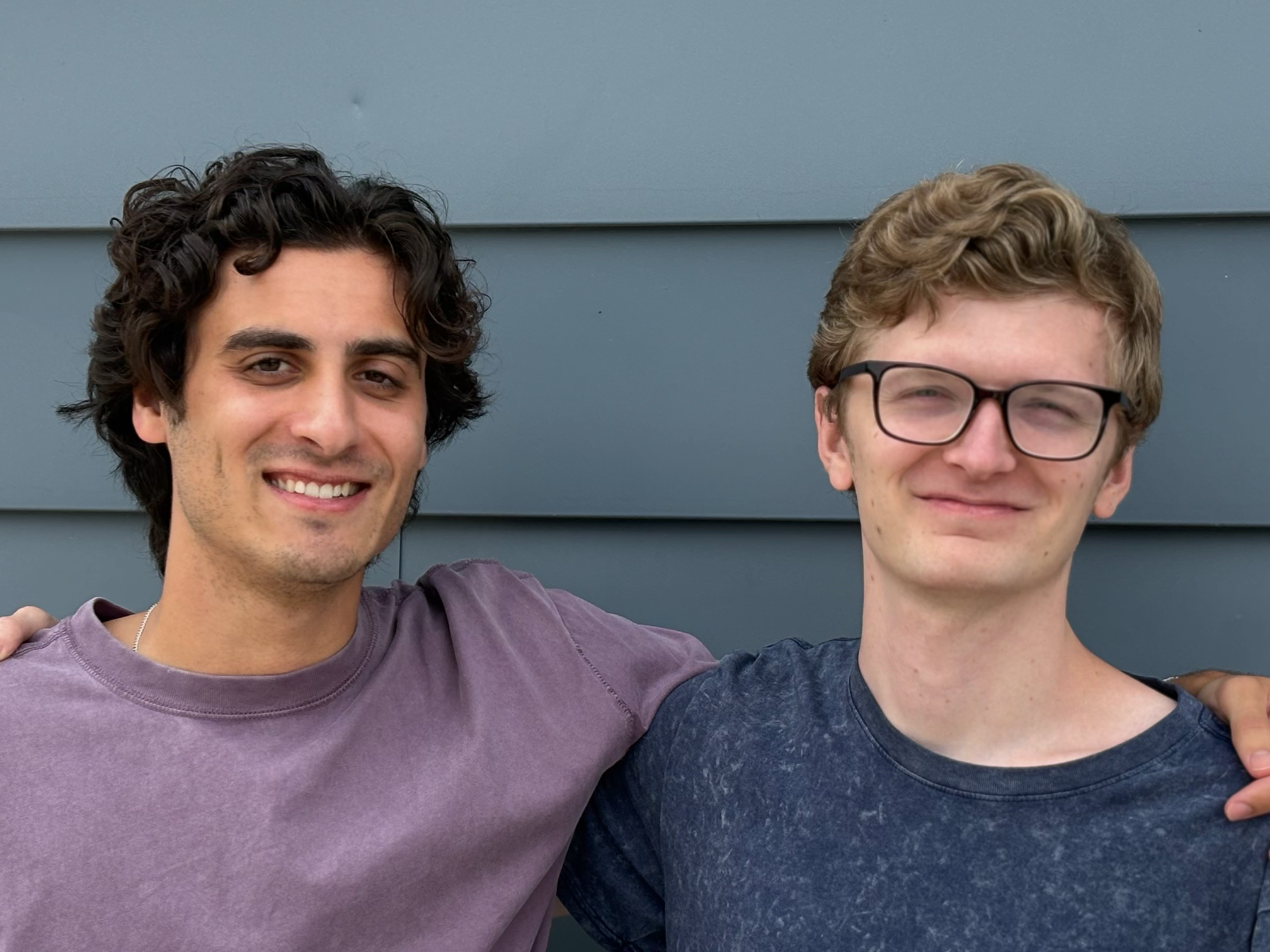 Nick Ponti and Andrew Hughes stand together, smiling arm in arm, in front of a blue wall.