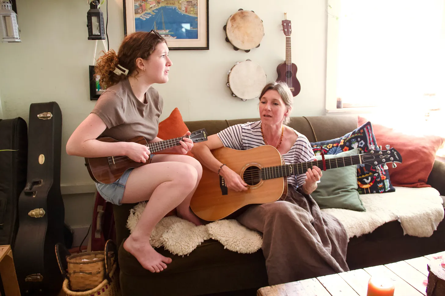 A girl playing ukelele sits next to a woman playing guitar on a couch. To their right lies a basket and instrument cases. Two tambourines, a ukelele, a painting, and other items hang on the wall behind them.