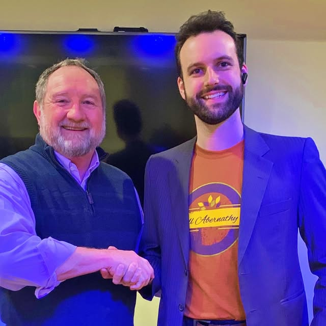 Bill shaking hands with a tour venue manager wearing a Bill Abernathy shirt under his jacket
