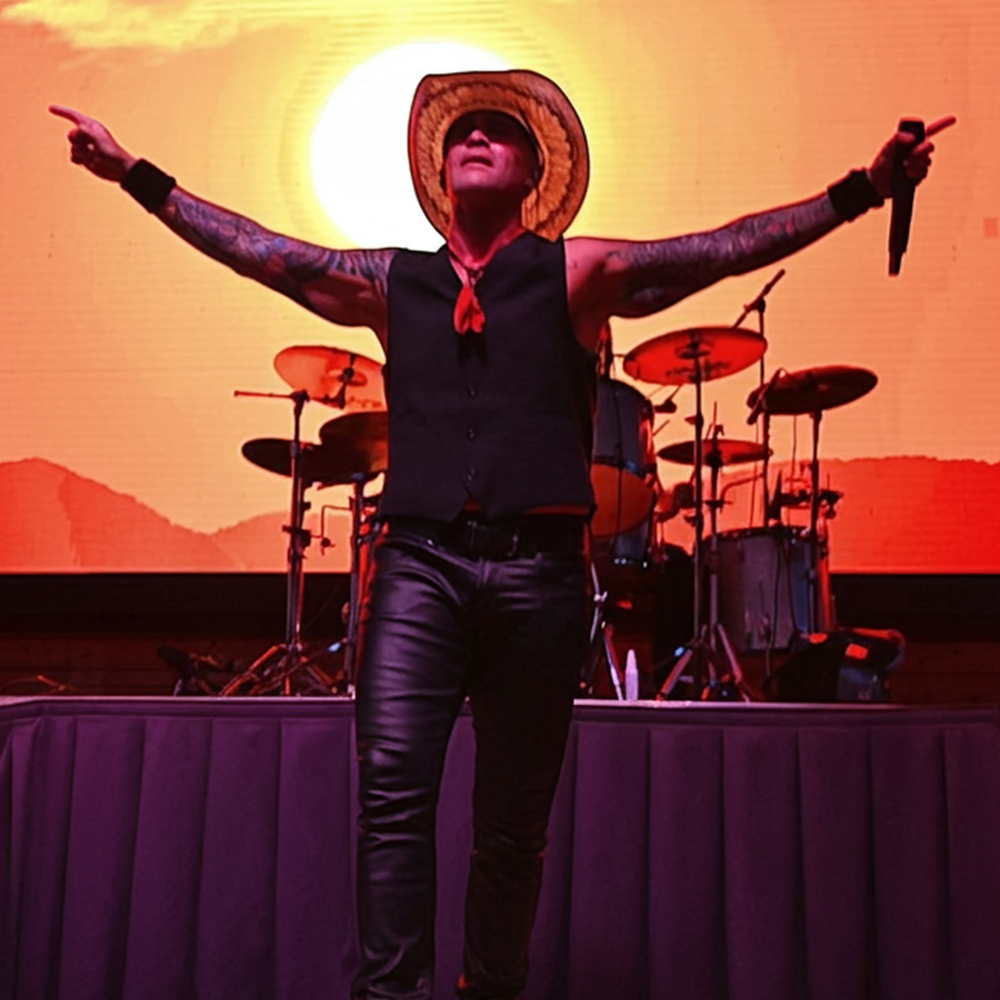 Pride U2 tribute's Jeff (Bono) in signature white hat and vest strikes iconic arms-raised pose while Mac (Edge) rocks the Gibson Explorer in red. Classic Vertigo-era staging with red lights.