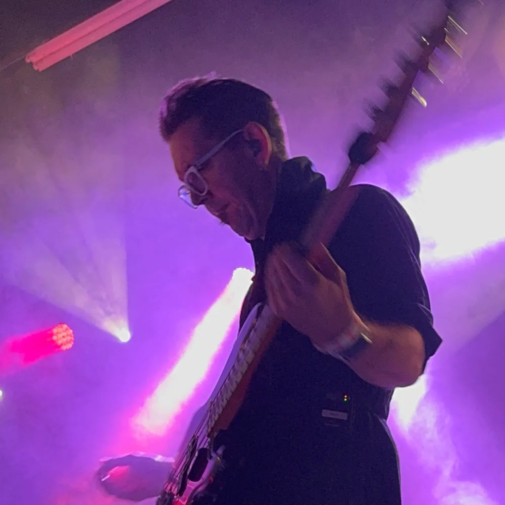 Jerko laying down bass lines on his precision bass, white-framed glasses and all-black attire, Pride's custom LED backdrop creating vibrant atmosphere. 