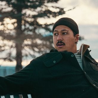 Photo of R'Know looking to the left while sitting outside wearing a black beanie and winter coat.