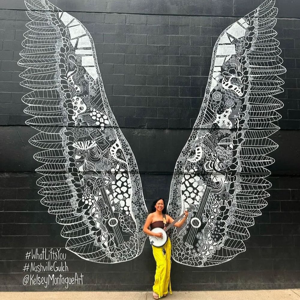 Silvia Passiflora, Southern Gothic Folk poet-songwriter, holding her banjo before the #WhatLiftsYou wings mural by Kelsey Montague in Nashville's Gulch neighborhood. 