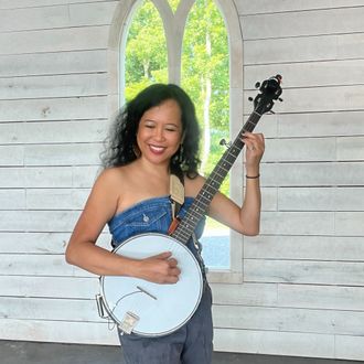 Silvia Passiflora, Southern Gothic Folk poet-songwriter, smiling and holding her banjo in a whitewashed wood chapel with an arched window framing summer greenery behind her.