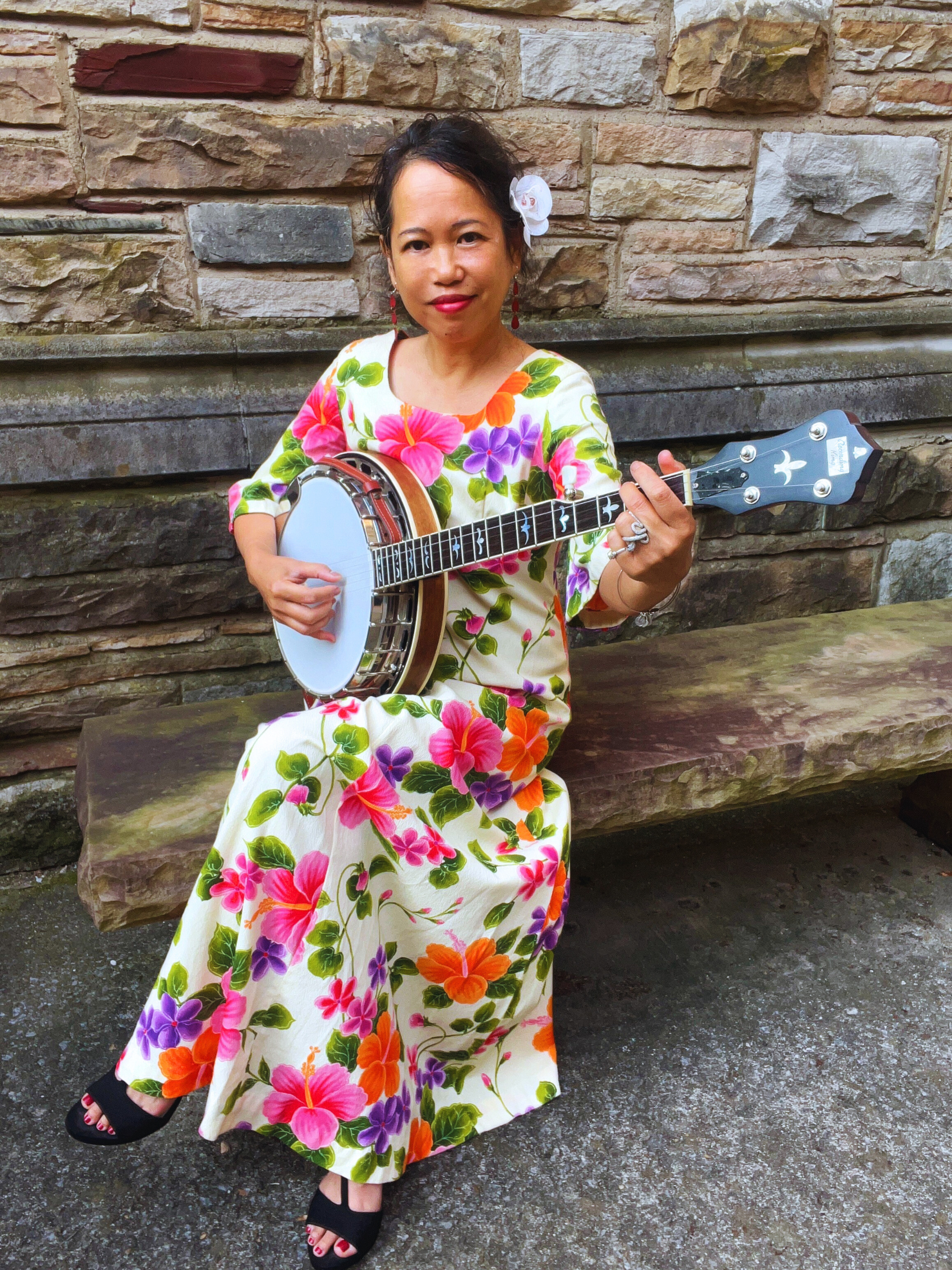 Silvia Passiflora, Southern Gothic Folk poet-songwriter playing a banjo, wearing a floral Hawaiiana gown and flower in her hair.