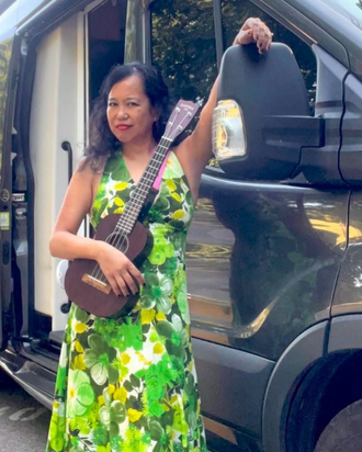 Silvia Passiflora, Southern Gothic Folk poet-songwriter, holding an ukulele beside her high-clearance tour van Hermie, wearing a green floral dress, ready for the road.