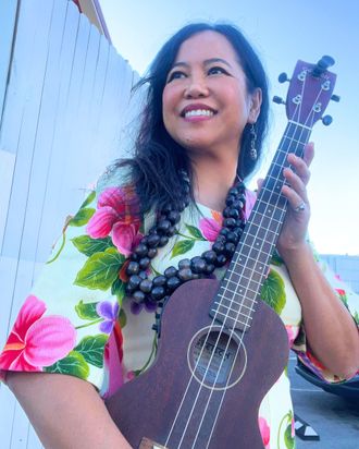 Silvia Passiflora, Southern Gothic Folk poet-songwriter, smiling upward while holding an ukulele outdoors, wearing a vibrant floral top and a bold black beaded necklace against a bright sky. 