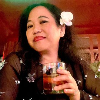 Silvia Passiflora, Southern Gothic Folk poet-songwriter, with a white flower in her hair, holding a cocktail glass and smiling warmly in a dimly lit intimate setting.