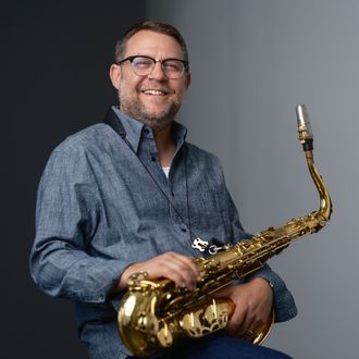 Michael Jenner holding tenor sax in his lap, smiling against a light blue-gray background