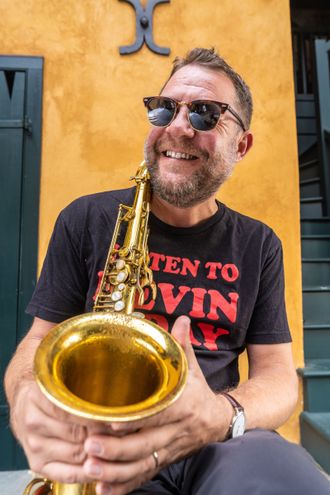 New Orleans Saxophonist Michael Jenner holding his saxophone in the French Quarter in front of a yellow wall, wearing a 'listen to Kevin O'Day' t-shirt