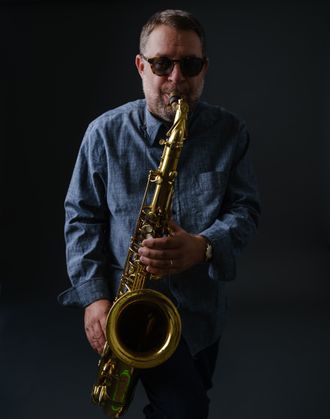 Michael Jenner playing tenor saxophone against a dark blue-gray background, wearing sunglasses and a collared linen shirt