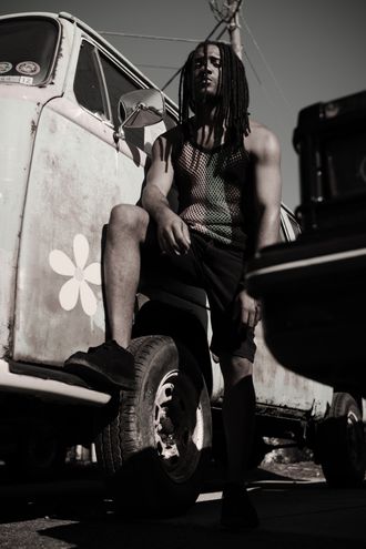 Jamaican artist EmE posing beside a vintage vehicle in an outdoor portrait junkyard 60's car vibe