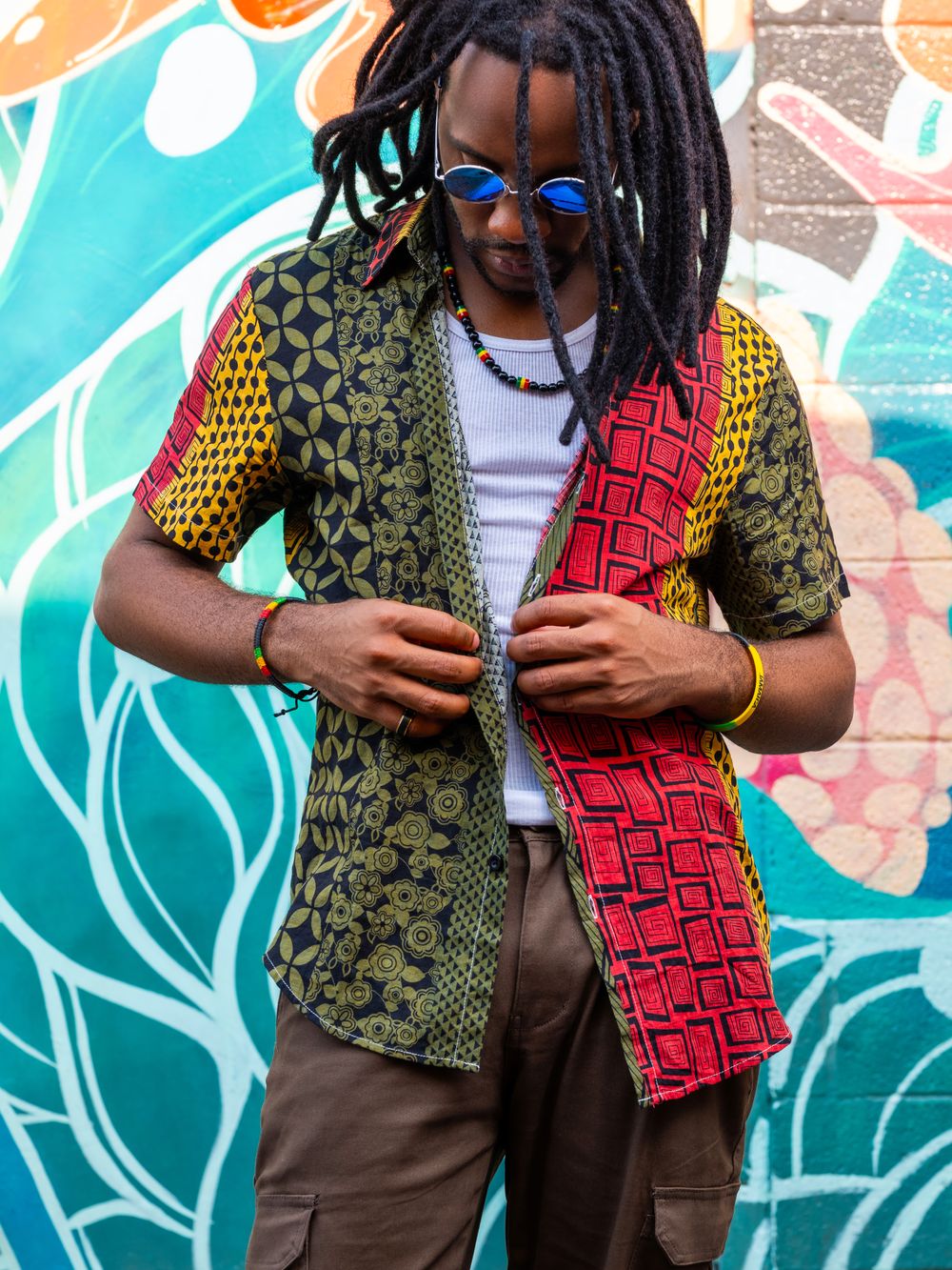 EmE, reggae-soul singer known for conscious lyrics and love songs, poses in front of a vibrant mural with locs, shades, and patterned rasta-inspired fabrics, merging genre fusion.