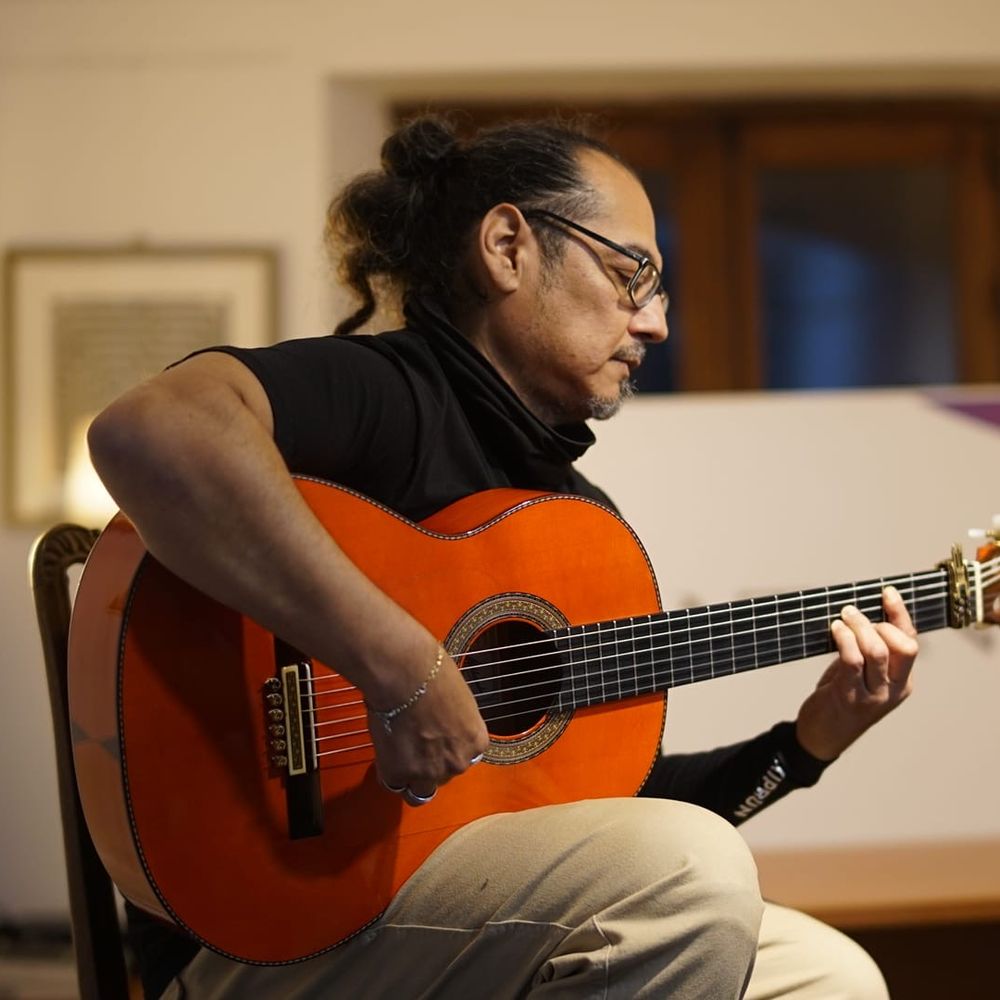 Shane Gonzalez strums an acoustic guitar, seated in front of a textured wall, immersed in his music.