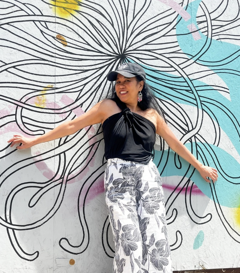Silvia Passiflora, Southern Gothic Folk poet-songwriter, in front of a mural vaguely resembling a flower. She is wearing a baseball cap, a black top and black and white floral print pants. 