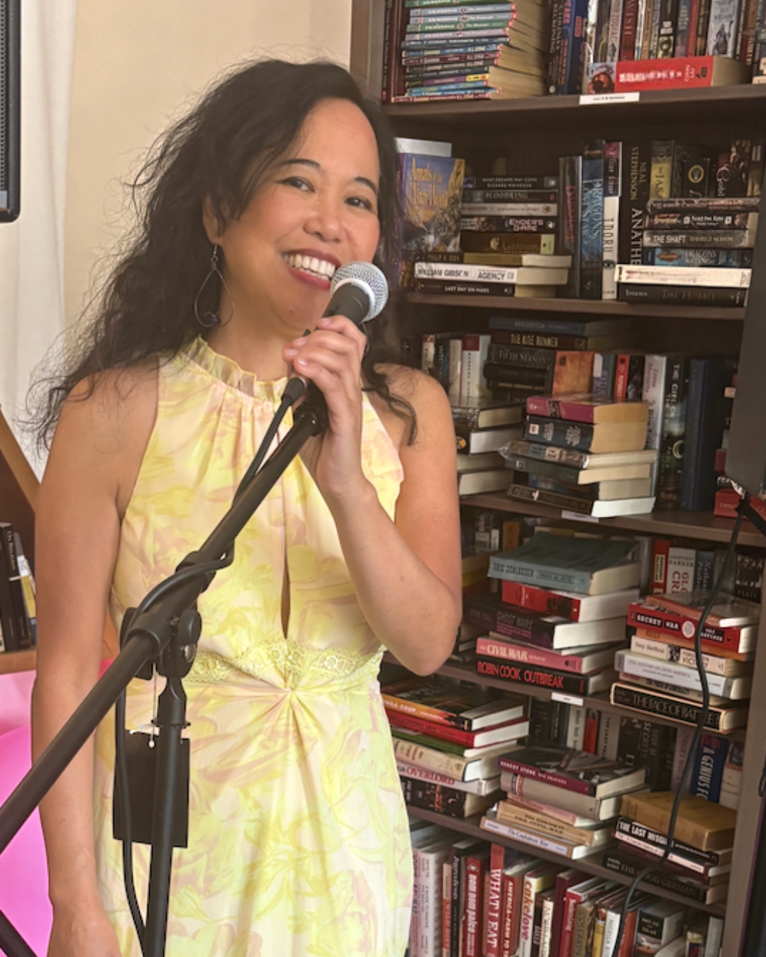 Silvia Passiflora, Southern Gothic Folk poet-songwriter, wearing a buttercup yellow gown behind a microphone stand in front of bookshelf at a bookstore, smiling at the viewer.