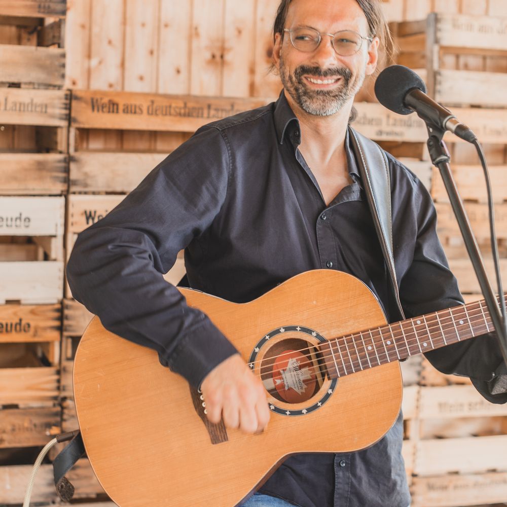 Live-Musiker Andy Wilsing mit Gitarre für Event und Hochzeit – Musiker im schwarzen Hemd vor rustikalem Hintergrund.