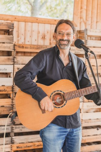 Sympathischer Hochzeitsmusiker Andy Wilsing mit Gitarre vor rustikaler Holzwand und Weinkisten – Live-Musik für Events in Karlsruhe.