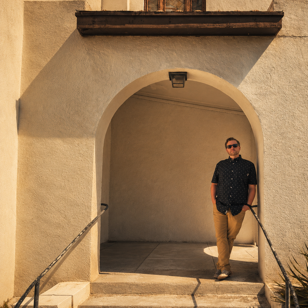 Musician Billy Lister in sunglasses leaning inside the arched entryway of an old stucco church in Marfa, Texas.