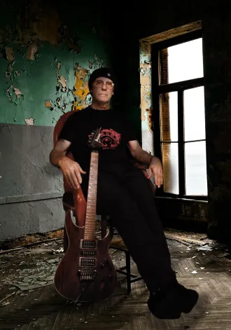 Andy is sitting in a chair in a room which has paint peeling off the walls and a heron bone parquet flooring. Andy is dressed in black and has his Ibanez guitar leaning against him. 