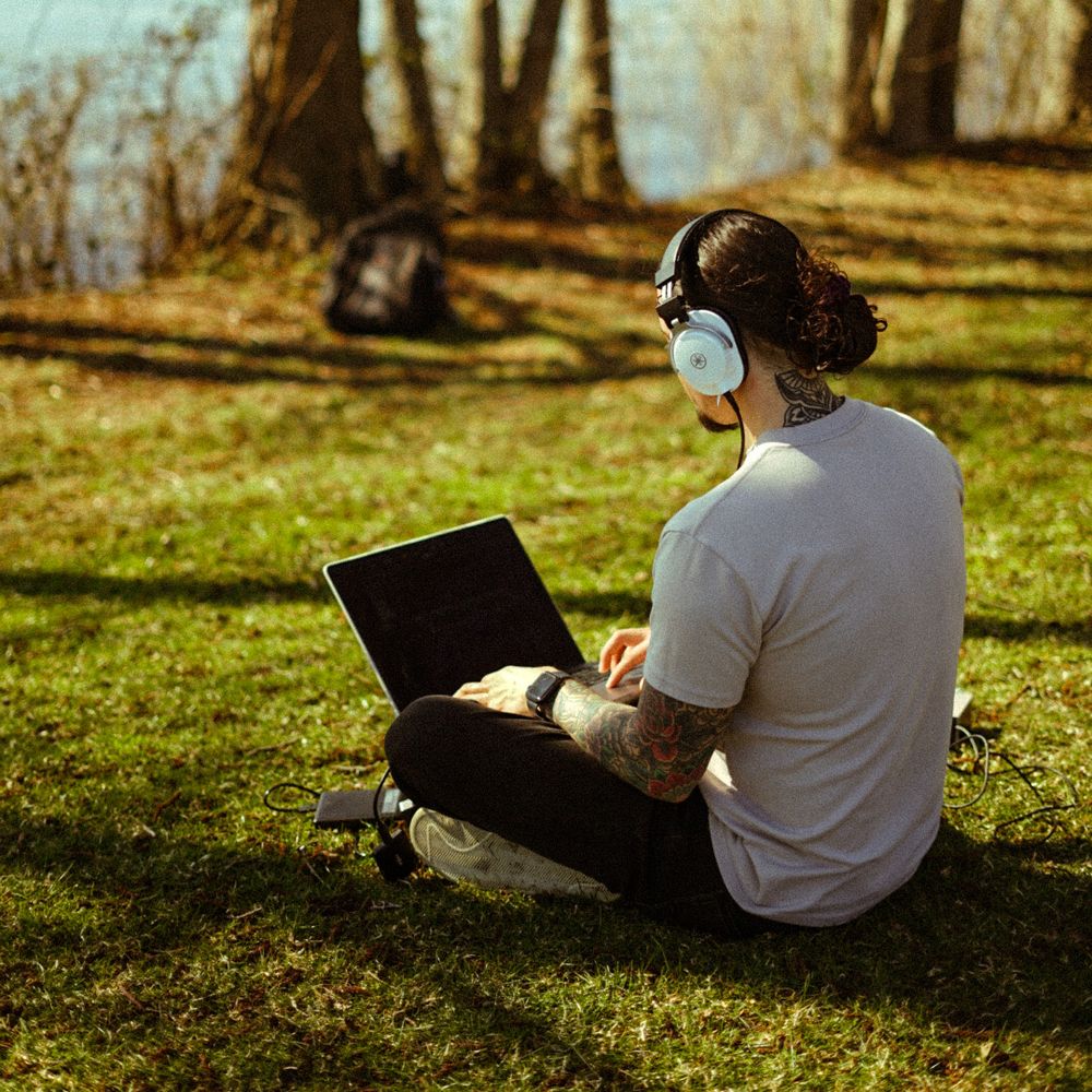 Waves the Creator producing music on a laptop outdoors by a lake while wearing headphones.