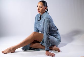 Madison Rose as Sade, seated on the ground with one leg pointed out, wearing iconic blue denim shirt and skirt