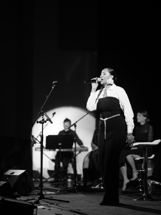 Black and white image of Madison Rose as Sade, playing live, singing into microphone with band in the background
