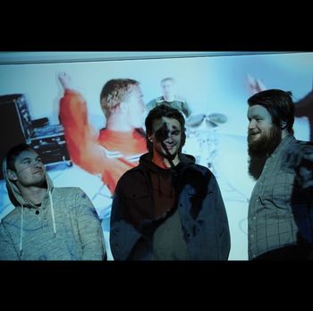 Adrian, DJ, and Troy posing for a photo for their original 3-piece Pop-Punk Band, "The Super Cedars" in 2019. Pictured on the projector screen behind them is a still from the music video "Dammit" by Blink-182.

