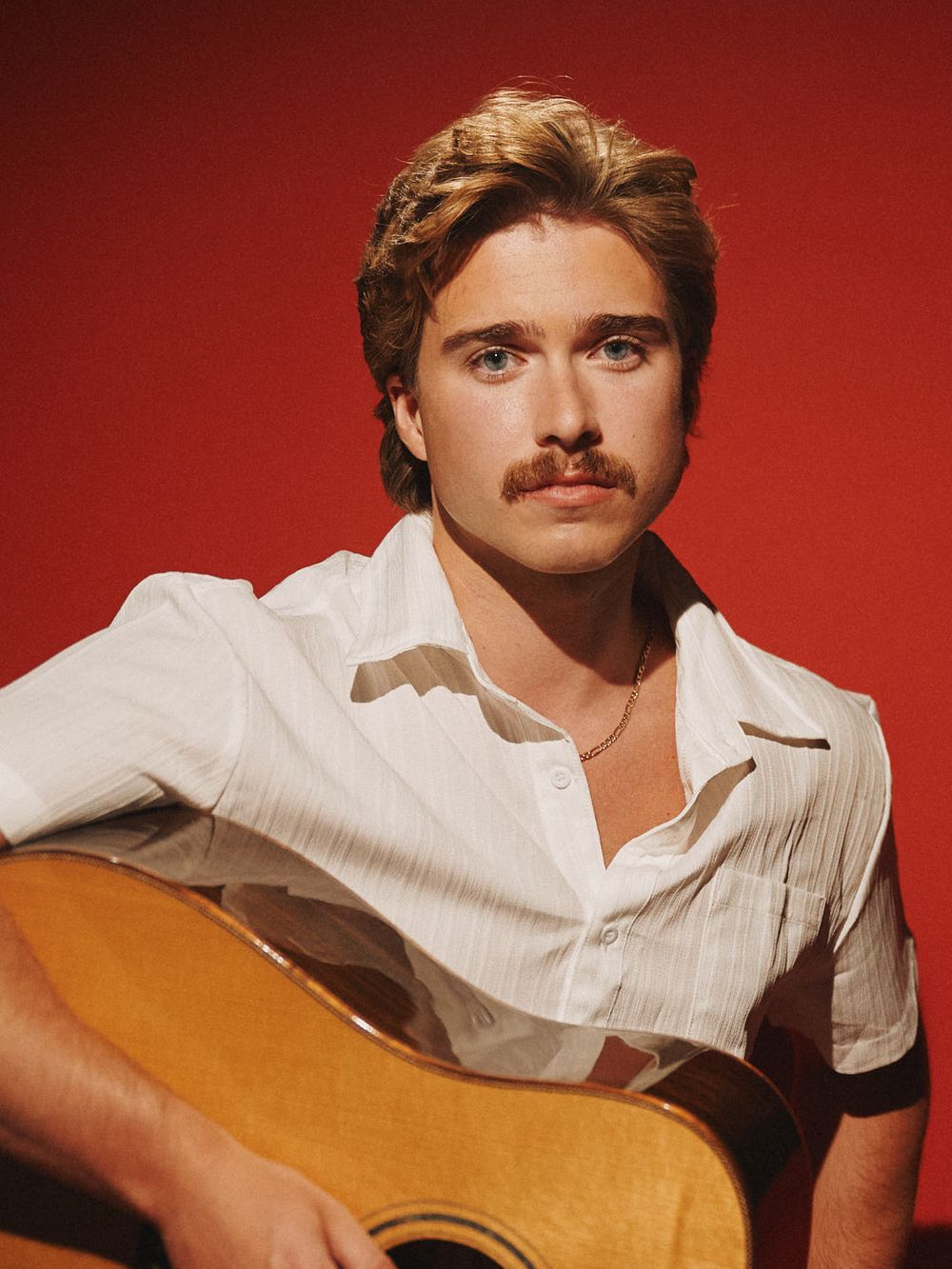 Tyson Ranger with an acoustic guitar against a red backdrop. Photo by Libby Danforth.