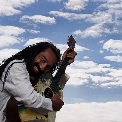 Adrian Xavier smiling with an acoustic guitar beneath a blue sky