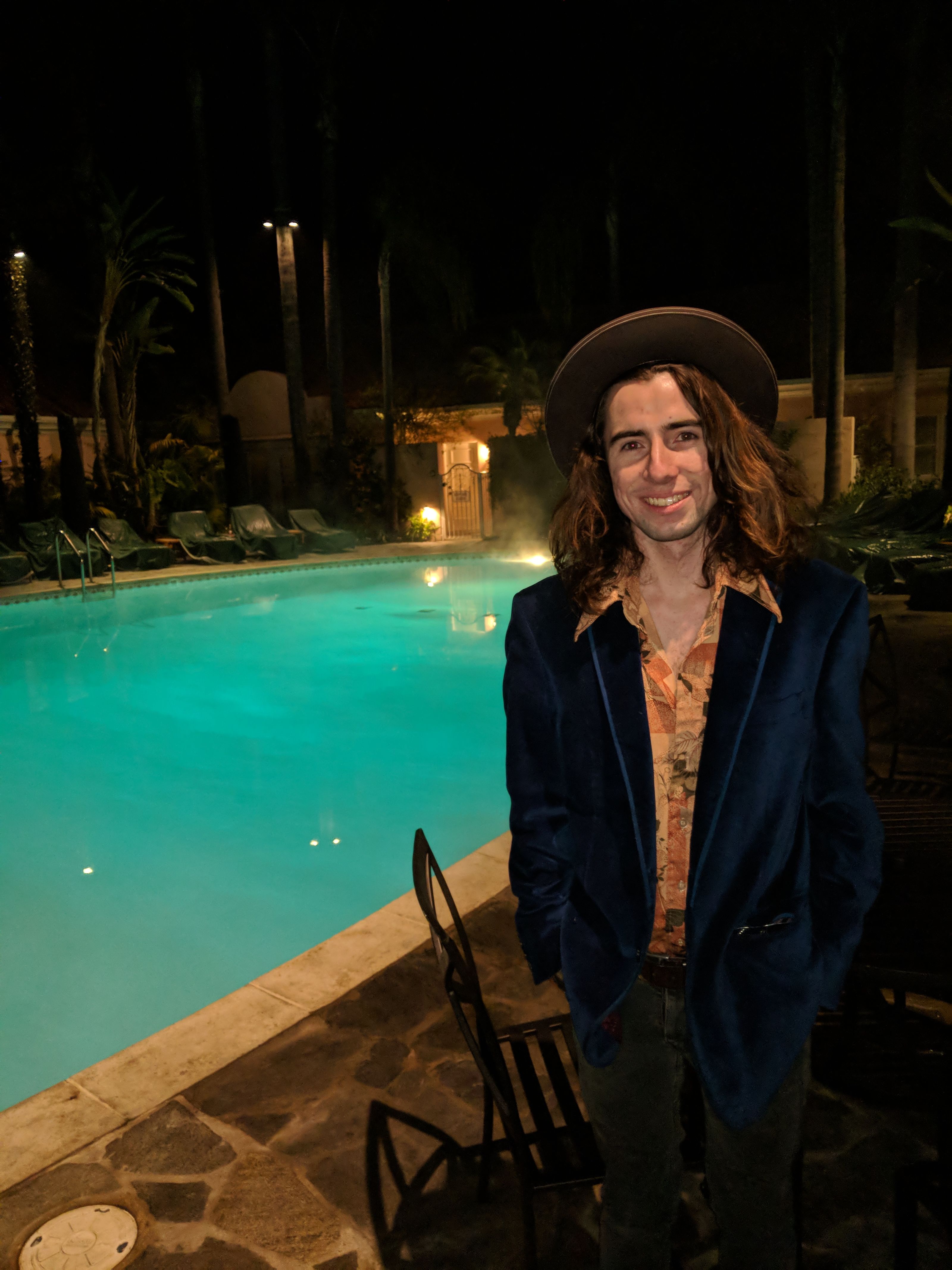 Harrison Flynn stands beside a lit swimming pool at night in a wide brim hat and dark jacket