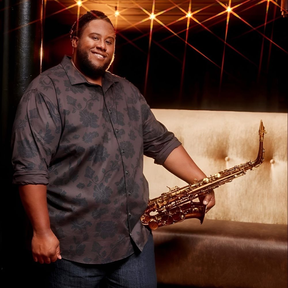 Grammy-winning saxophonist David TV Barnes smiling and holding his saxophone in a patterned shirt under warm stage lights