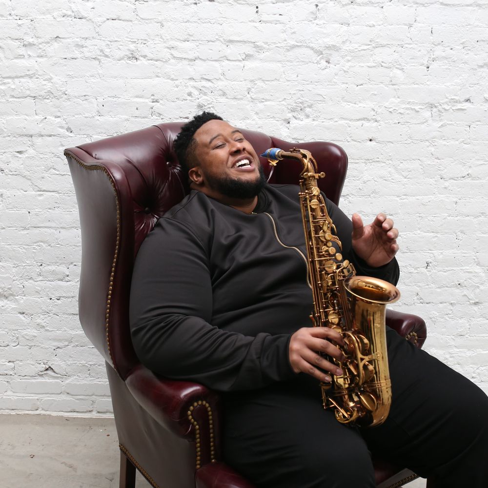 Grammy-winning saxophonist David TV Barnes laughing in a leather chair, holding his saxophone against a white brick wall