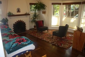 An inviting room with two chairs, a fireplace, a bow window, and a massage table with a colorful cover of aqua and large magnet hibiscus flowers. 