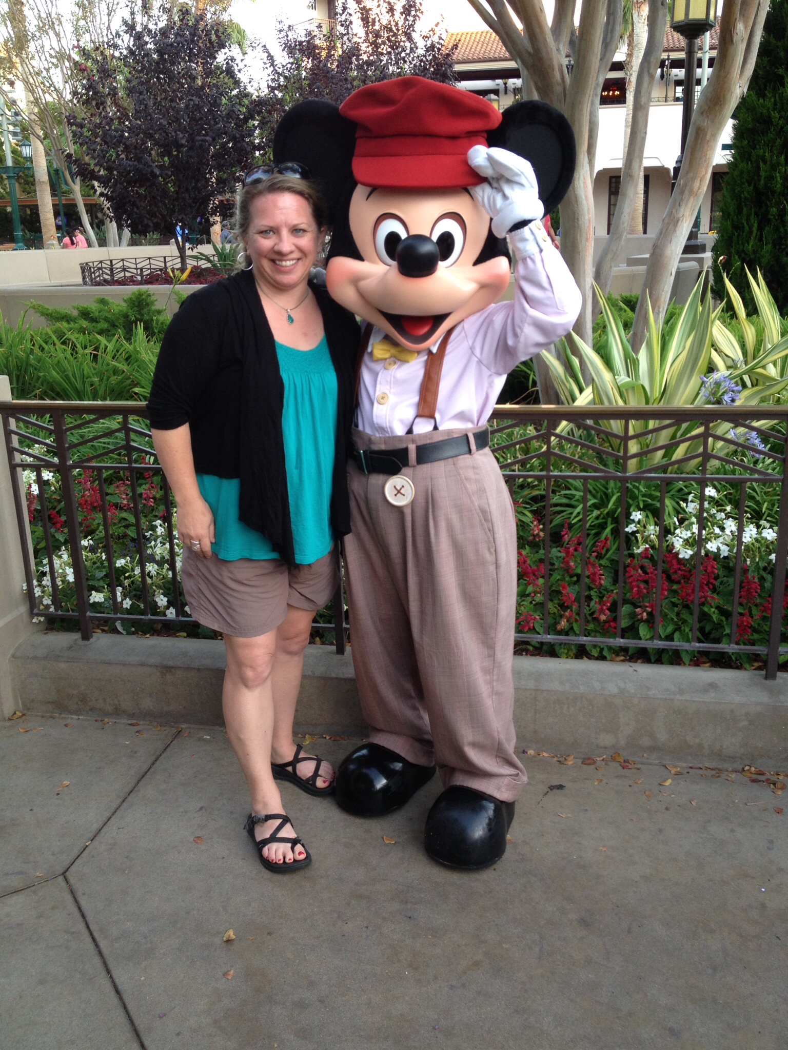 Blond woman in green shirt and brown shorts with a black sweater standing next to and hugging Mickey Mouse dressed in khaki pants, white shirt and suspenders and a red hat